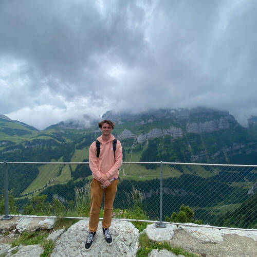 Grant Gordon standing on a mountain side in Switzerland in July of 2024 while wearing a peach sweatshirt and khaki pants with a cloudy sky.